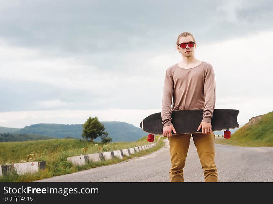 A stylish young man standing along a winding mountain road with a skate or longboard in his hands the evening after sunset. The concept of youth sports and travel hobbies.