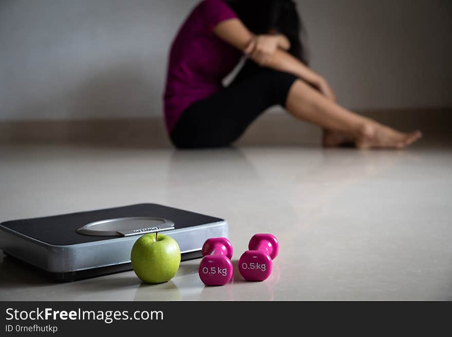 Close up weighing Scale in front of upset woman with measuring tape, pink dumbbell and green apple. Healthy lifestyle, food and sport concept.