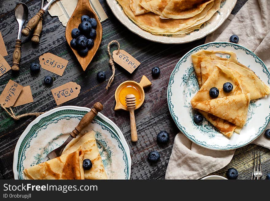 Overhead shot of thin yummy crepes with honey, blueberries on three vintage plates with green ornament on grey wooden table with honey, berries, forks, napkin. breakfast still life