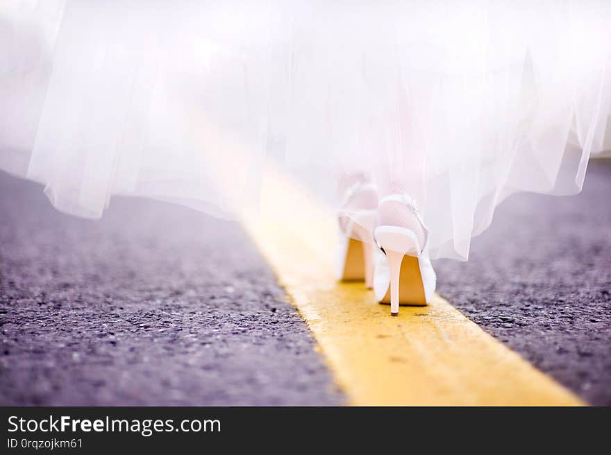 Closeup of the legs of the bride in shoes. She goes exactly along the yellow line of the road marking along the highway, asphalt is visible and the bride is moving away from the camera. Closeup of the legs of the bride in shoes. She goes exactly along the yellow line of the road marking along the highway, asphalt is visible and the bride is moving away from the camera