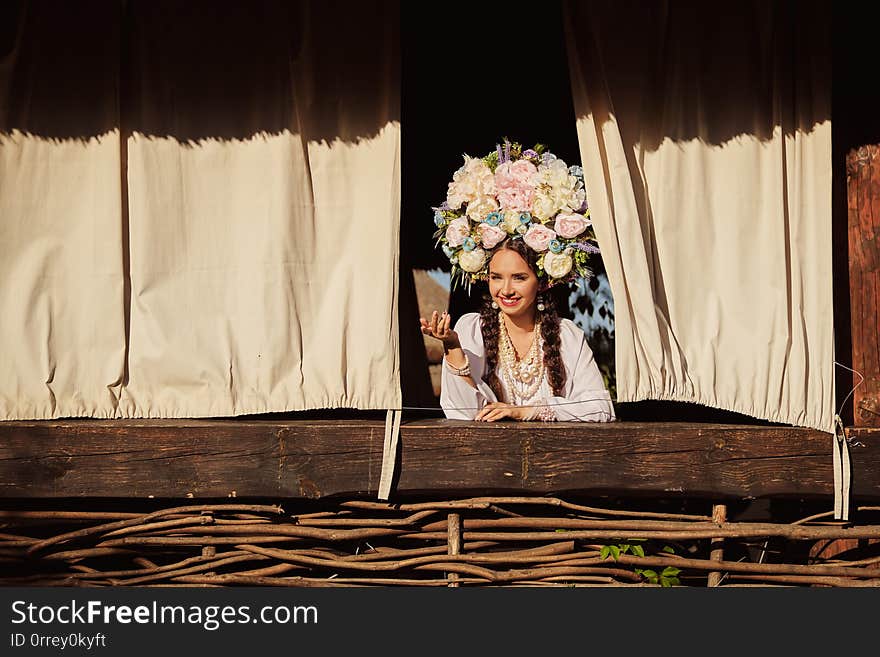 Beautiful brunette female with a long dark hair, in a white ukrainian authentic national costume and a wreath of colorful flowers is smiling and looking out of the window. Beautiful brunette female with a long dark hair, in a white ukrainian authentic national costume and a wreath of colorful flowers is smiling and looking out of the window.