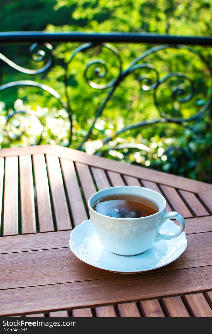 Single white ceramic cup of tea on a saucer on wooden table against picturesque and natural green foliage background. Single white ceramic cup of tea on a saucer on wooden table against picturesque and natural green foliage background.