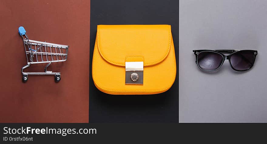 Minimalistic Shopaholic Concept. Yellow leather bag, mini shopping trolley sunglasses on paper background. Women&#x27;s fashion accessories. Top view