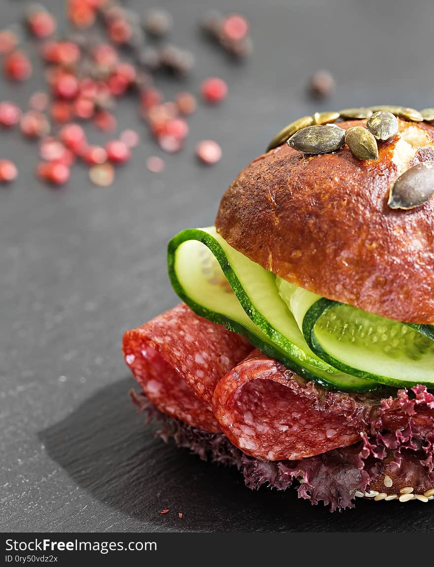 Burger with salami, lettuce and cucumber. Bun with sesame seeds and pumpkin seeds. Quick breakfast. Black stone background. Close-up