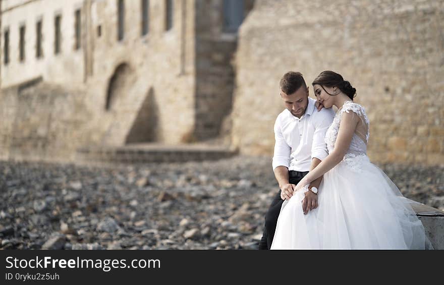 Close-up of beautiful fairytale newlywed couple sitting on the bench and hugging at rocky beach near old medieval castle