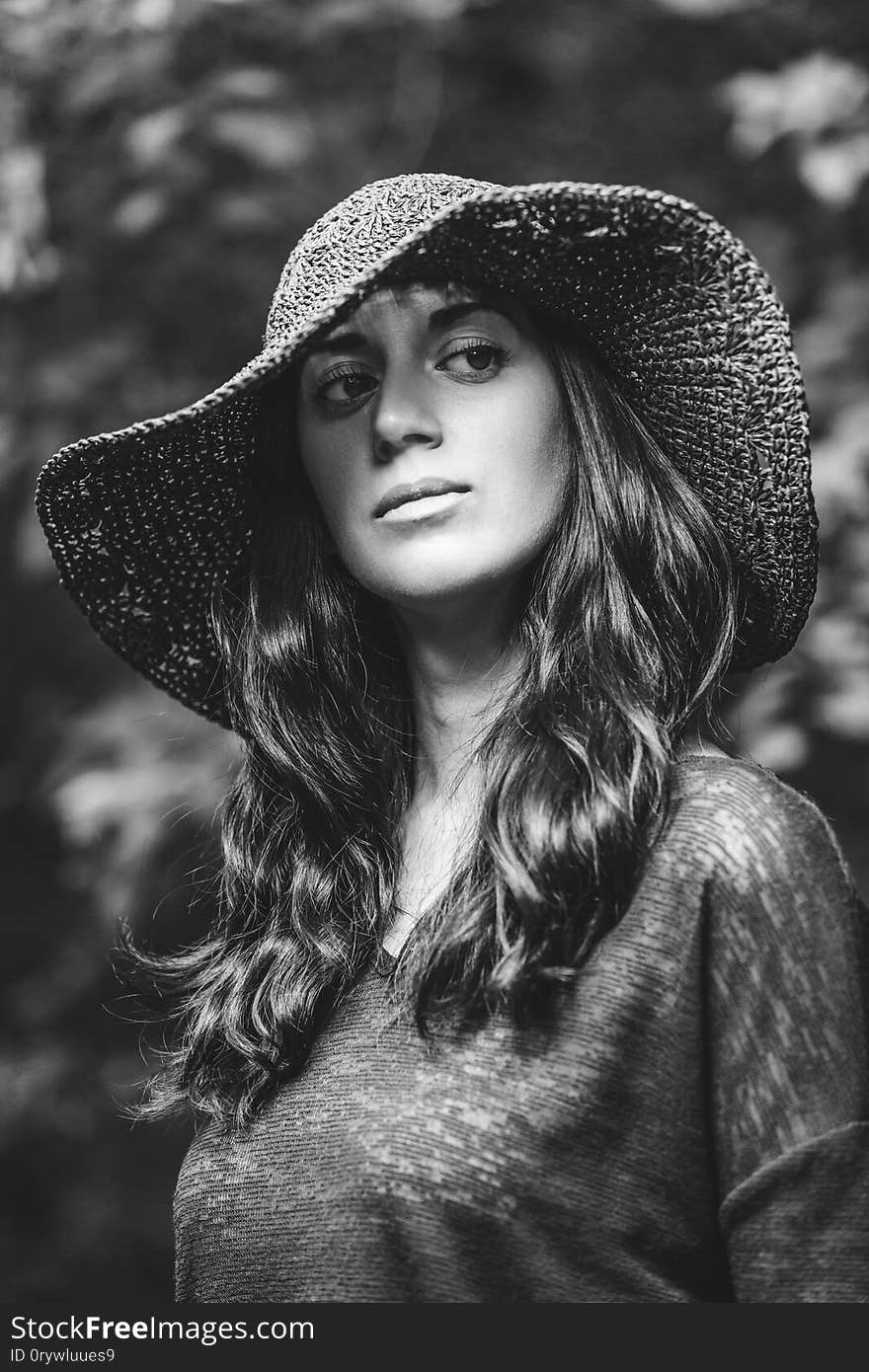 Close up portrait of a beautiful girl in blue dress and black hat standing on background of green leaves. Sunlight breaks through the hat and falls on her face. Vertically framed shot