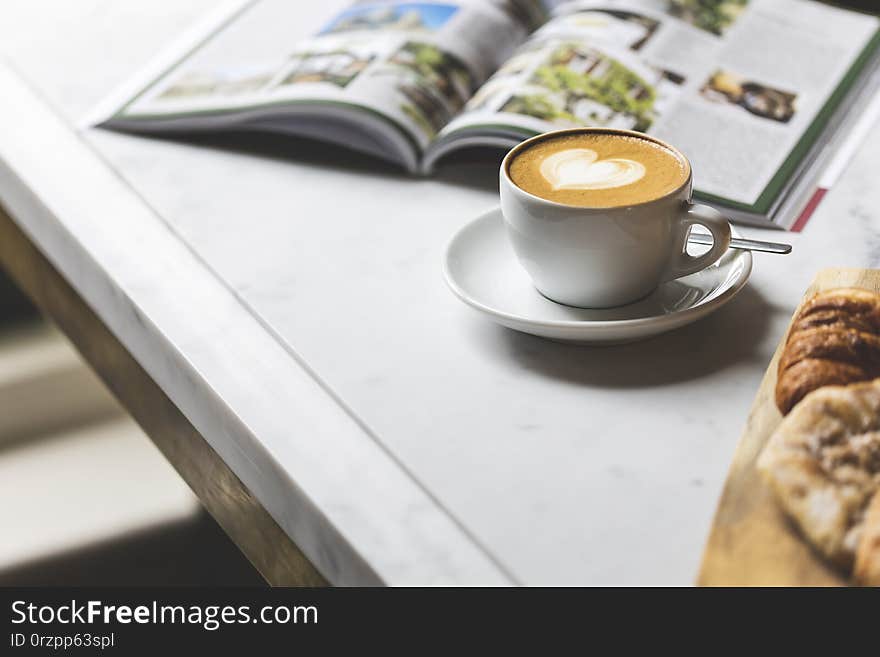 Cup Of Cappuccino With A Cute Saucer Heart, A Croissant, And A Magazine On The White Table