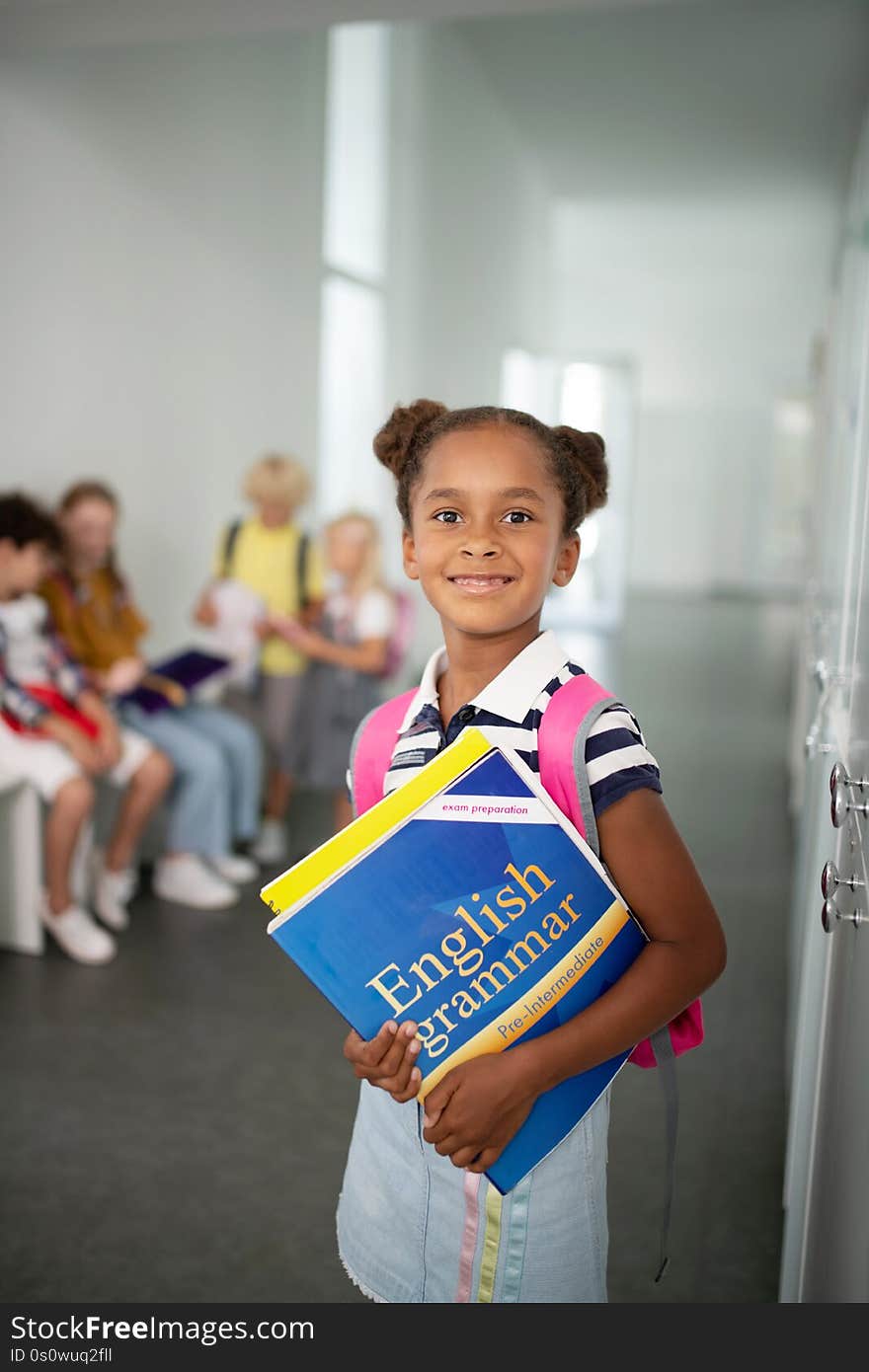 Cute dark-skinned girl holding books while standing at school