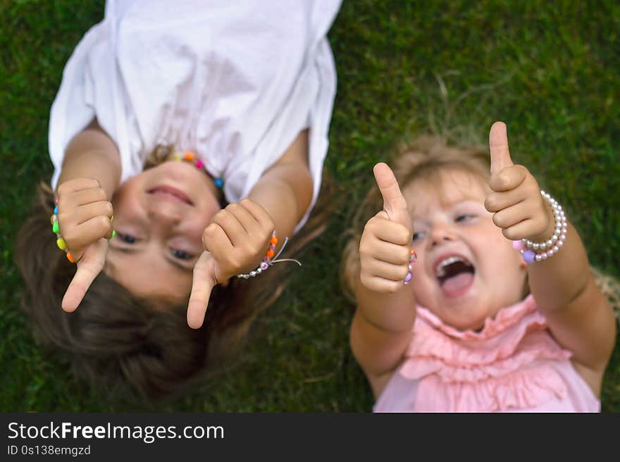 Two Little Girls Laying On The Grass, Laughing And Showing Thumbs Up