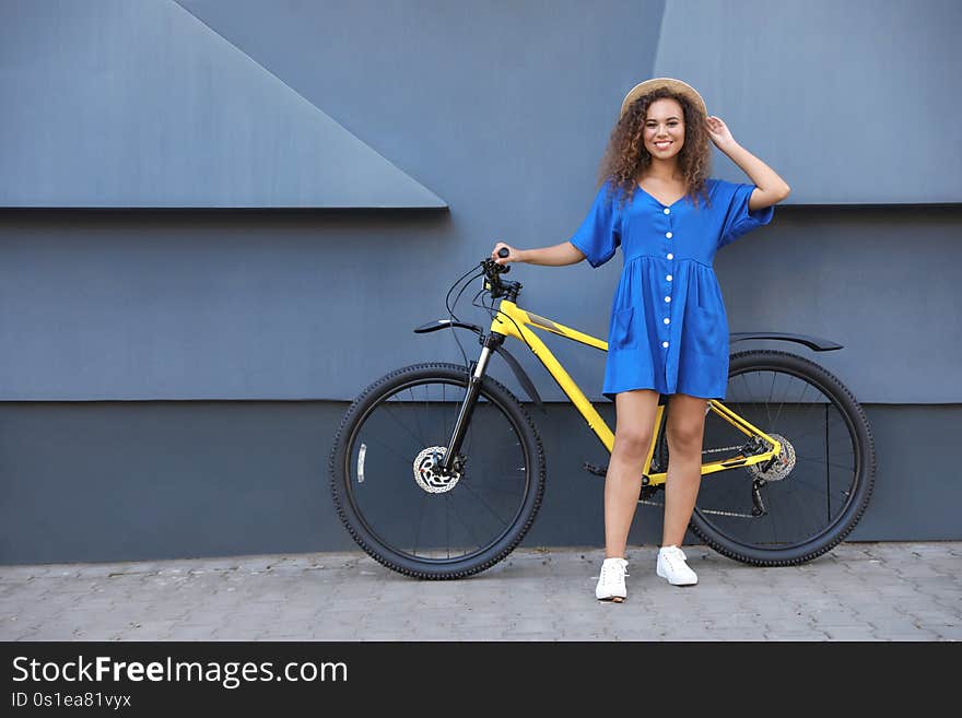 Beautiful young African-American woman with bicycle near grey wall on city street. Space for text