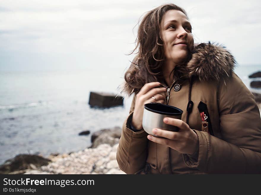 Smiling beautiful young woman standing on sea coast during cloudy weather with traveler cup in hands