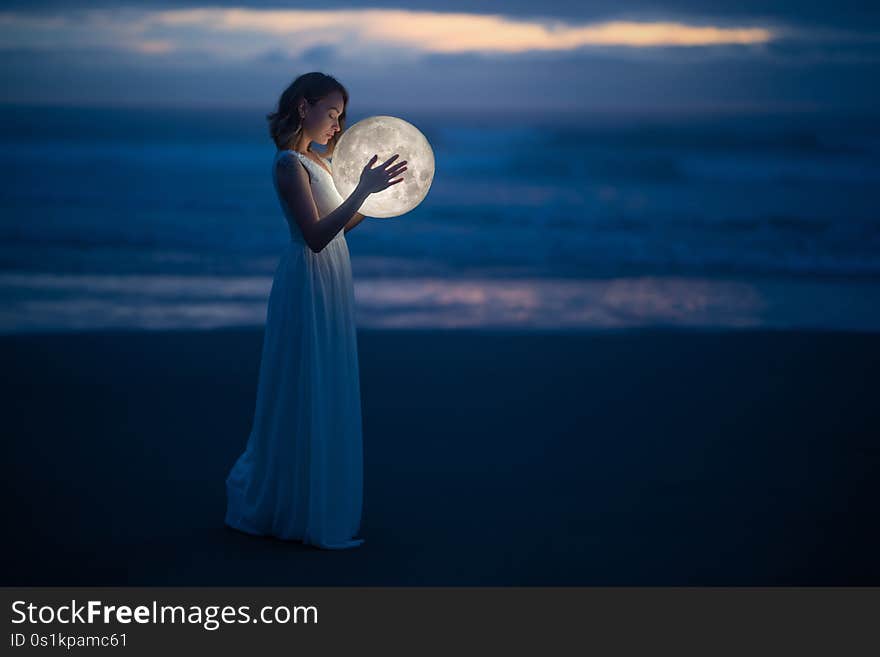 Tender image of a girl. Female magic. Beautiful attractive girl in full growth on a night beach with sand hugs the moon, art photo