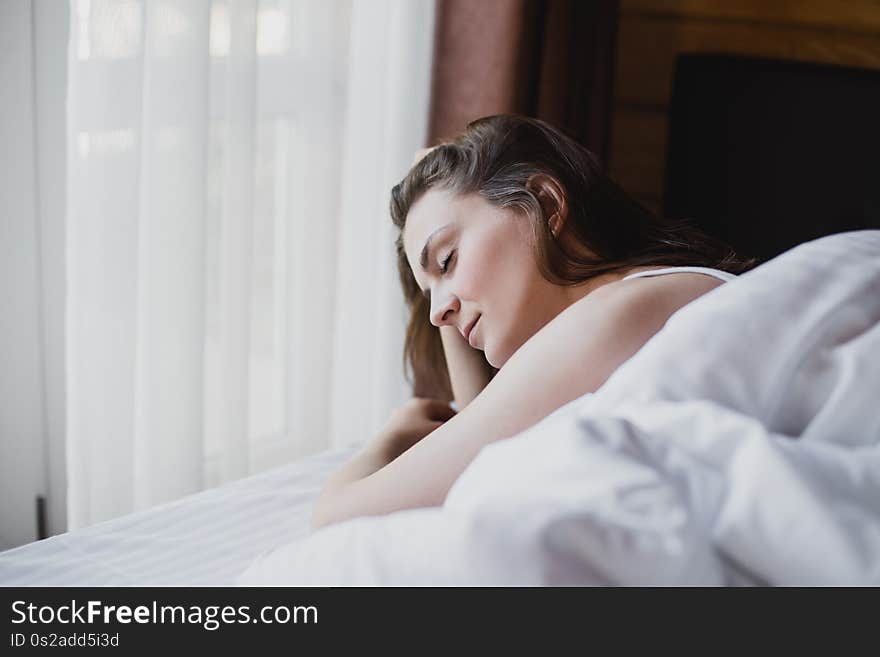 Beautiful young woman having good morning in bed with clean white linens in a cozy wooden house. She is looking at window and having dreamy mood. Beautiful young woman having good morning in bed with clean white linens in a cozy wooden house. She is looking at window and having dreamy mood.