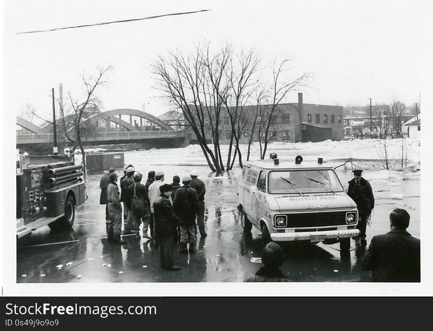 A photograph showing police, firefighters and city workers standing next to the Moira River during the 1981 flood in Belleville, Ontario. The Bell Shirt Company and the Lower Bridge can be seen in the background. This photo was taken by Elaine Preston. A photograph showing police, firefighters and city workers standing next to the Moira River during the 1981 flood in Belleville, Ontario. The Bell Shirt Company and the Lower Bridge can be seen in the background. This photo was taken by Elaine Preston.