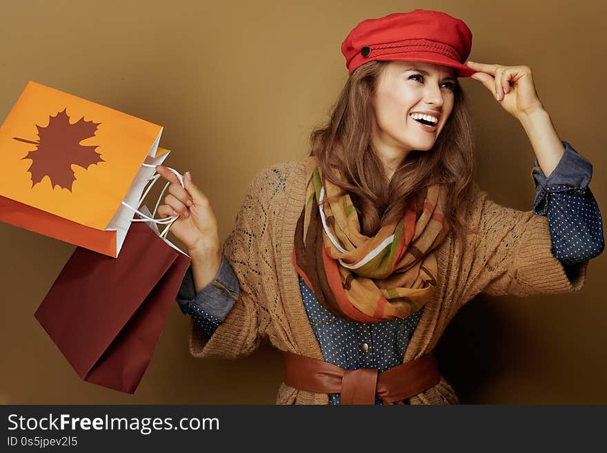 Smiling modern middle age woman in red cap, scarf, jeans shirt and cardigan with autumn shopping bags looking aside  on brown background. Smiling modern middle age woman in red cap, scarf, jeans shirt and cardigan with autumn shopping bags looking aside  on brown background