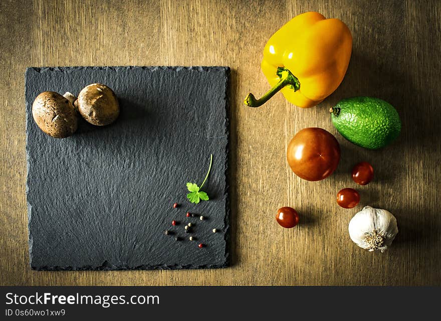 A vegetable composition with pepper, tomato, avocado, garlic and mushrooms on a black chopping board