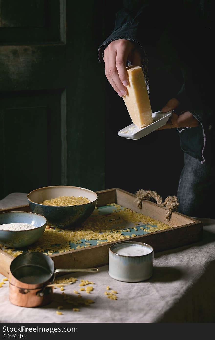 Young man in black grate cheese. Raw uncooked italian pasta coquillettes for cooking dinner with parmesan, cream and semolina flour on wooden tray on kitchen linen table cloth. Dark rustic style.