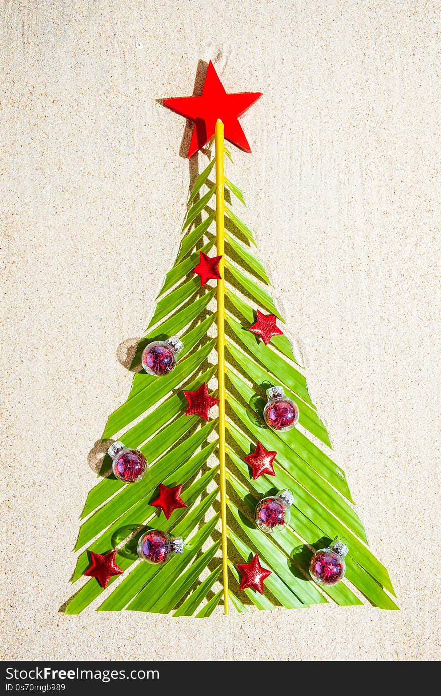 Christmas tree made of green palm thee brunch with small red festive decorations on sand, top view, vertical composition.