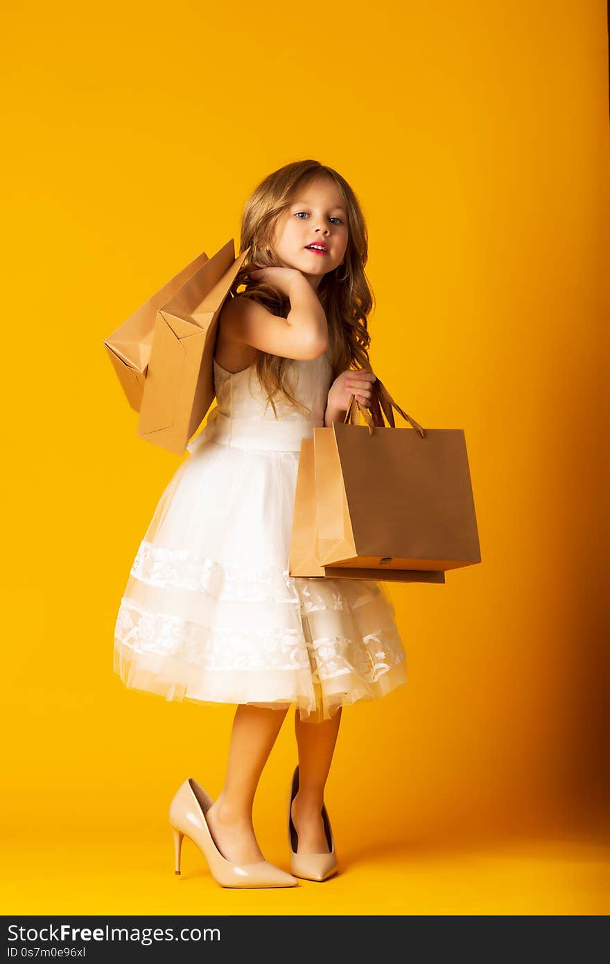 Funny Child Girl Fashionista In Big Mother`s Shoes On Yellow Background With Packages In Hand.