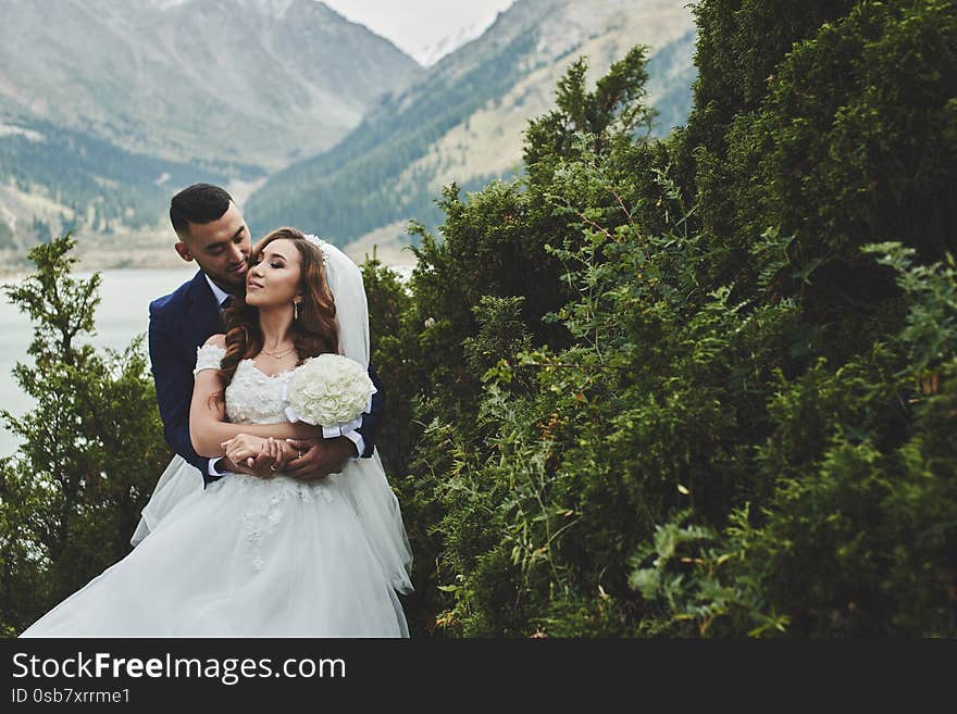 Beautiful wedding photo on mountain lake. Happy Asian couple in love, bride in white dress and groom in suit are photographed agai