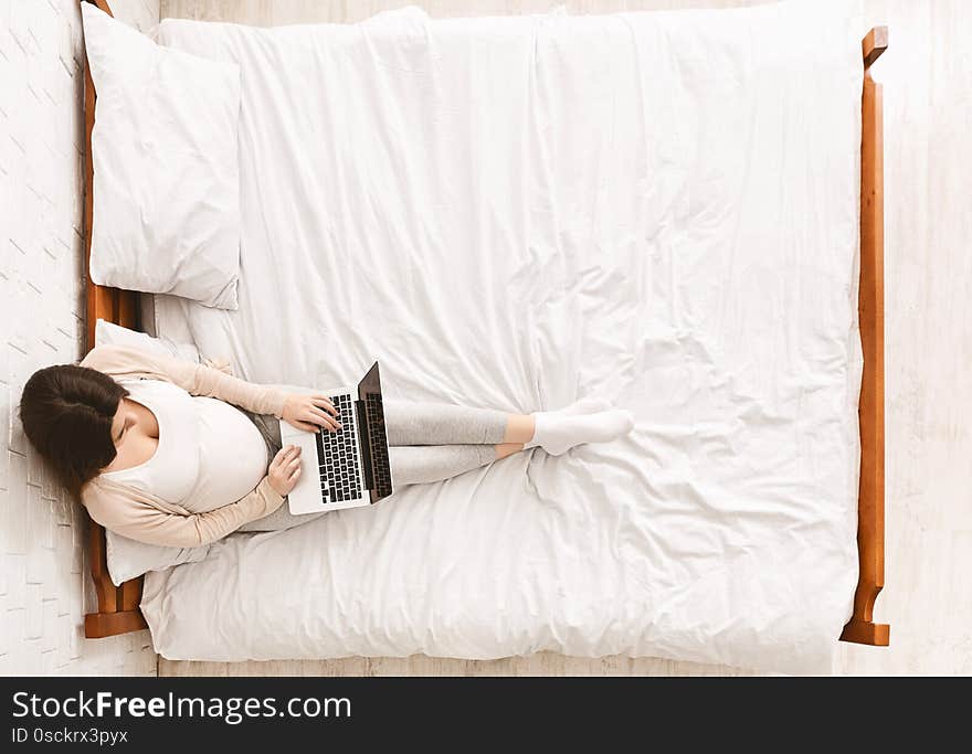 Pregnant woman working on laptop, sitting on bed at home, top view with empty space