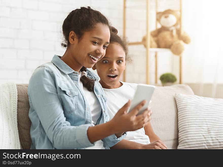 Shocking mobile app. African girl showing her little sister new mobile application, sitting at home, copy space. Shocking mobile app. African girl showing her little sister new mobile application, sitting at home, copy space