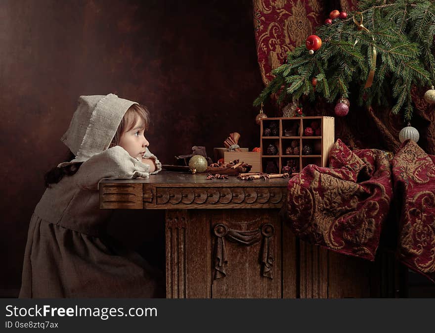 Little girl in an vintage linen dress dreaming near the table with sweets. Chocolates and spices on the table under the Christmas tree. Genre portrait in retro style. Little girl in an vintage linen dress dreaming near the table with sweets. Chocolates and spices on the table under the Christmas tree. Genre portrait in retro style