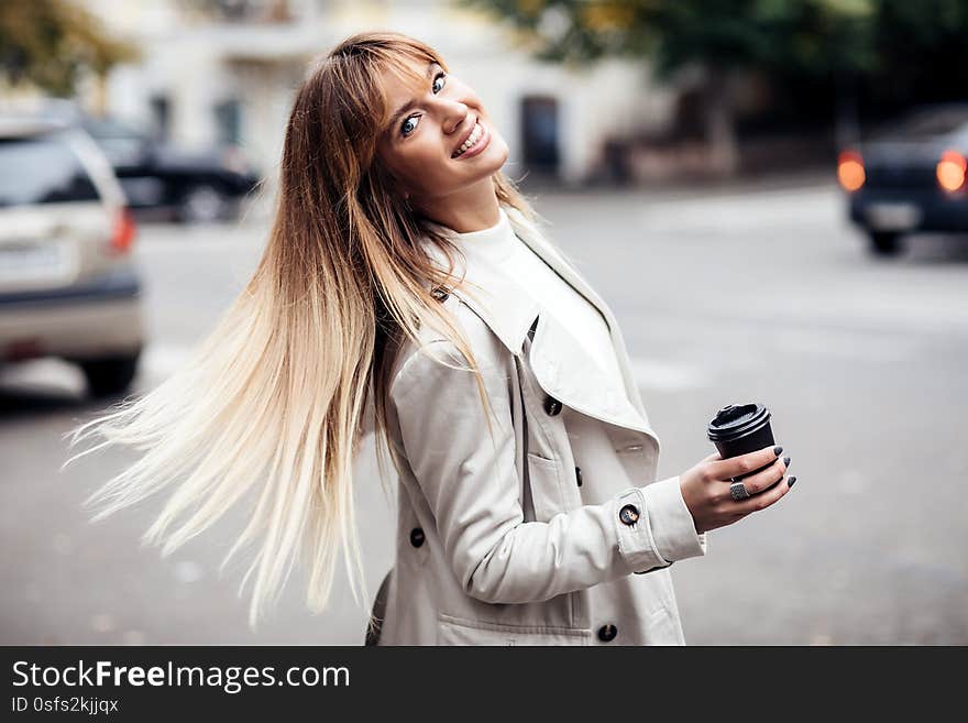 Stylish beautiful woman blonde in beige trench coat holds coffee to go. portrait of smiling girl . Street fashion concept.Dynamically young girl walks down the street.Hair fluttering in the wind. Stylish beautiful woman blonde in beige trench coat holds coffee to go. portrait of smiling girl . Street fashion concept.Dynamically young girl walks down the street.Hair fluttering in the wind