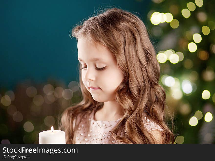 Portrait of acute long-haired little girl in dress on background of  lights.Little girl holding burning candle. Christmas, New Year and birthday celebration concept. Winter holidays. Copy space