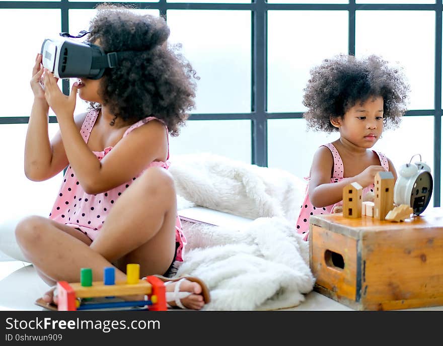 Soft blur image of one African mixed race girl enjoy with VR glasses while the other girl as main focus subject  play with some toys and they sit on floor.