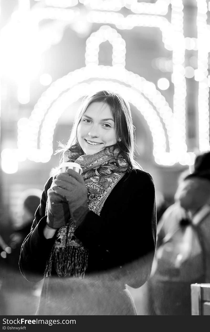 Black and white photo of young girl with glass in her hands on street in evening against background of burning garlands