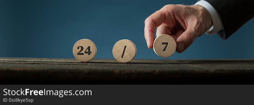 Wide view image of a hand of a businessman assembling a 24/7 sign with wooden cut circles. Over blue background. Wide view image of a hand of a businessman assembling a 24/7 sign with wooden cut circles. Over blue background