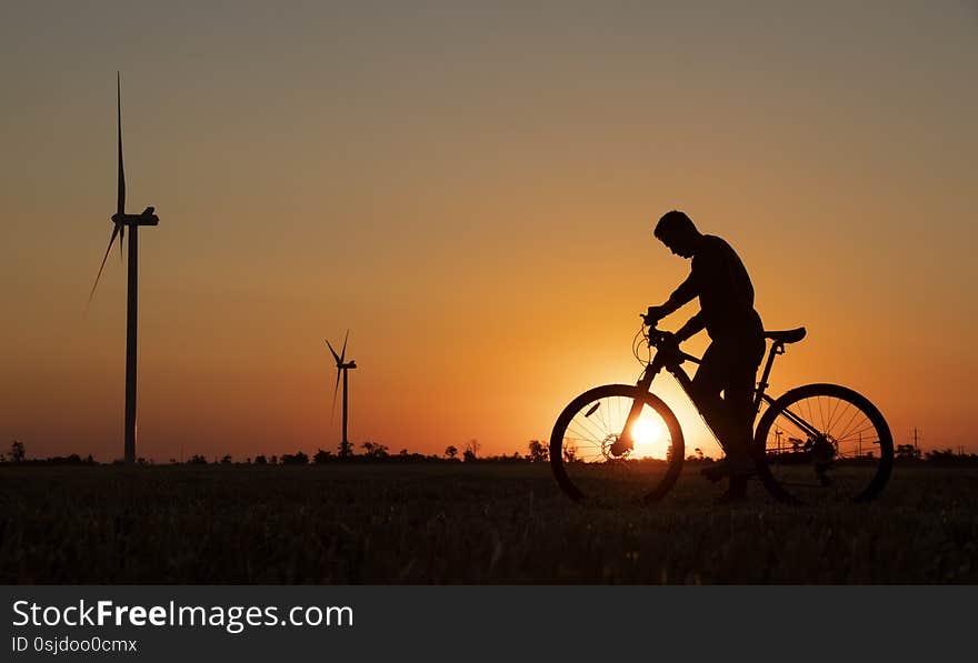 A cyclist with a camera stands in a field during the sunset against the background of an orange sky with the sun and windmills.