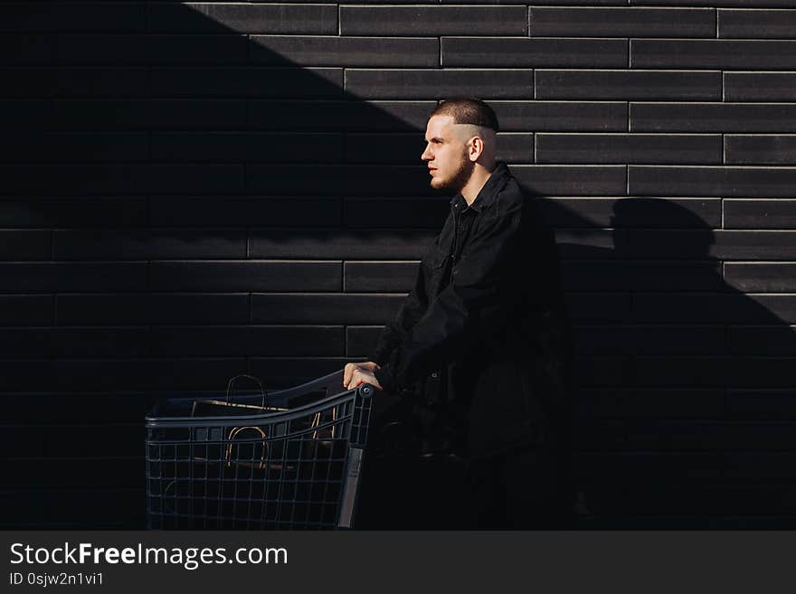 Young man is wearing black clothes and posing next to the black wall. He is holding a trolley with bags in it after shopping on Black Friday. Young man is wearing black clothes and posing next to the black wall. He is holding a trolley with bags in it after shopping on Black Friday