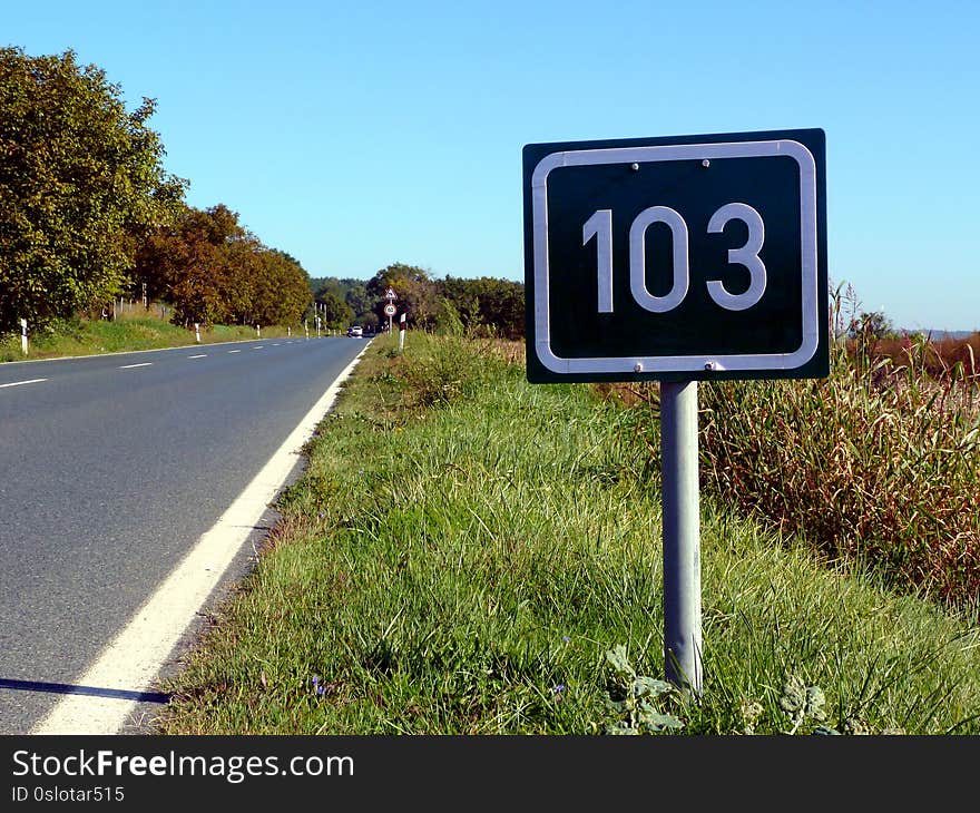 Deep green metal plate mileage sign. white lettering on road side. gray asphalt paved highway beyond. blue sky and trees. travel and transportation concept. summer scene. diminishing perspective. Deep green metal plate mileage sign. white lettering on road side. gray asphalt paved highway beyond. blue sky and trees. travel and transportation concept. summer scene. diminishing perspective