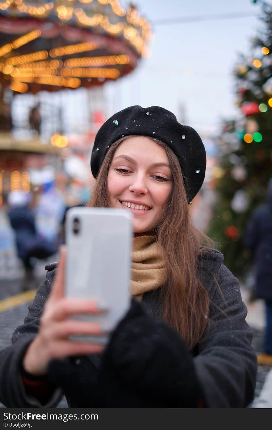 Beautiful young woman taking a selfie with christmas tree behind her with mobile phone camera - Pretty girl enjoying