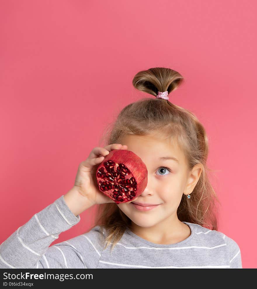 Smiling positive little girl in a gray sweater, holding a half pomegranate, closing one chapter, looking up at the advertisement, in high spirits, healthy eating, children`s menu, models on a pink studio background. Smiling positive little girl in a gray sweater, holding a half pomegranate, closing one chapter, looking up at the advertisement, in high spirits, healthy eating, children`s menu, models on a pink studio background