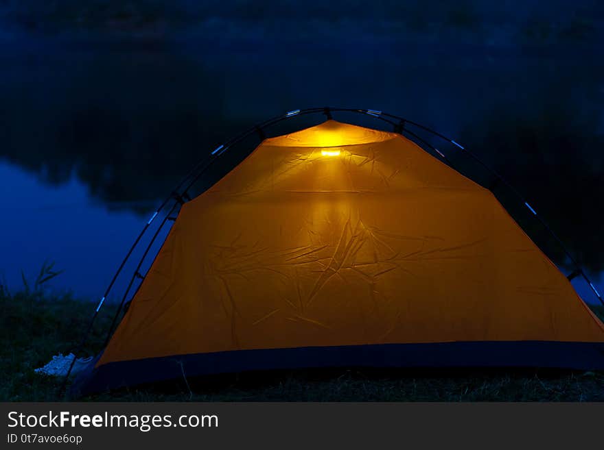 An orange tent at night stands on the shore and glows inside.