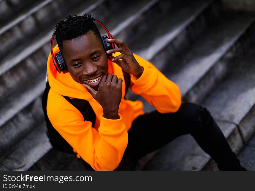 Happy african-american man in stylish orange hoodie sweatshirt in wireless headphones listening music and sitting on stairs. copy space. Happy african-american man in stylish orange hoodie sweatshirt in wireless headphones listening music and sitting on stairs. copy space