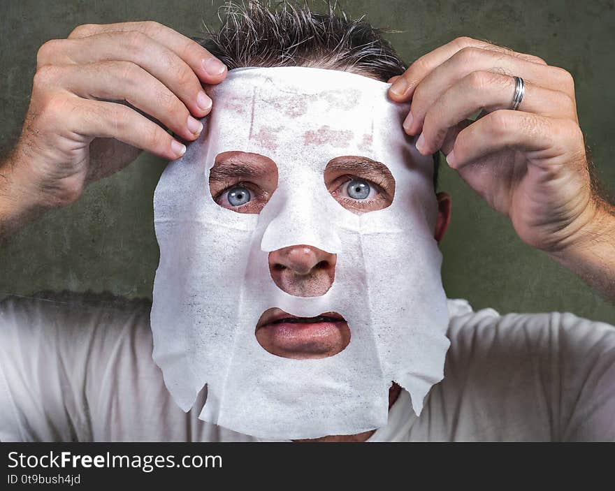 Lifestyle isolated background portrait of young weird and funny man at home trying using paper facial mask cleansing applying anti aging beauty treatment in hilarious face expression