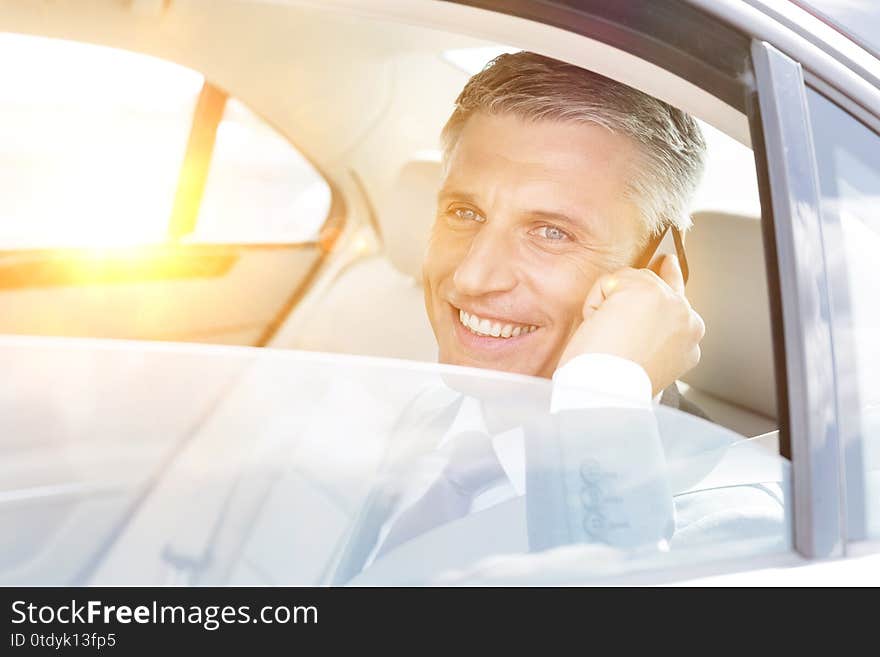 Smiling mature businessman talking on smartphone while looking through window in car with yellow lens flare in background