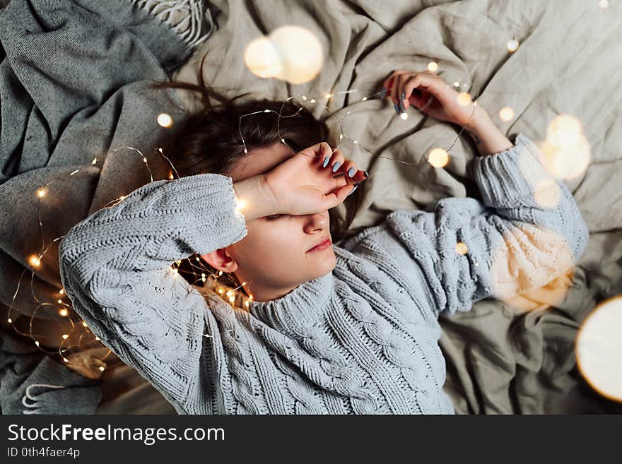 Young white woman lying in bed with fairy Christmas lights and bokeh around, shallow selective focus