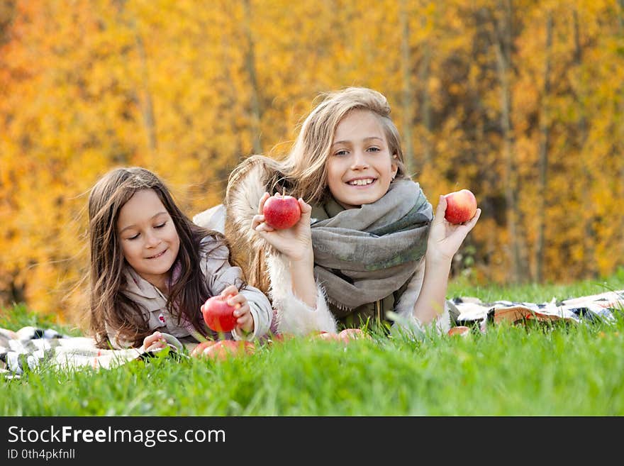 Beautiful caucasian big and little sisters lie on plaid, play with red apples on background of green grass of golden trees of autumn forest. Concept family weekend outdoors, people, lifestyle