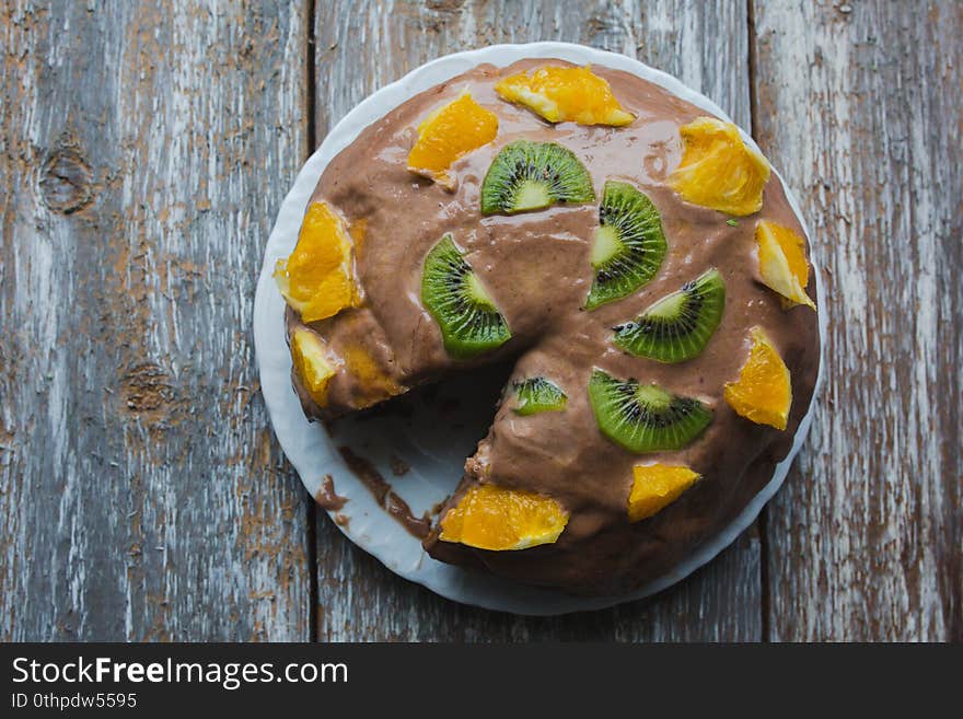 Homemade sponge cake with chocolate cacao ganache frosting. Slices of kiwi and orange on the dessert. Baked pastry in the kitchen on the old vintage wooden background.