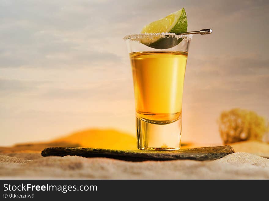 Gold Tequila In A Glass With Salt And A Slice Of Lime On A Skewer Stands On A Stone Against A Beautiful Sunset In The