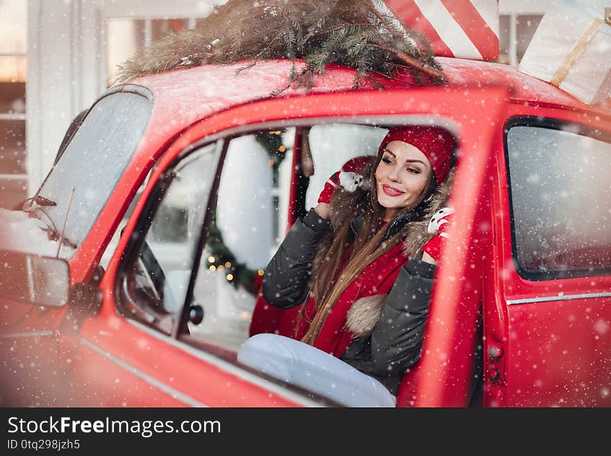 Pleasant fashionable winter girl smiling posing at red vintage car surrounded by snowflakes