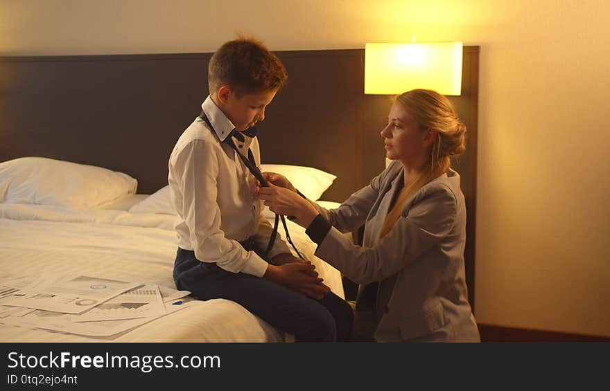 Woman in suit put a tie on little boy, who sit on the bed in hotel