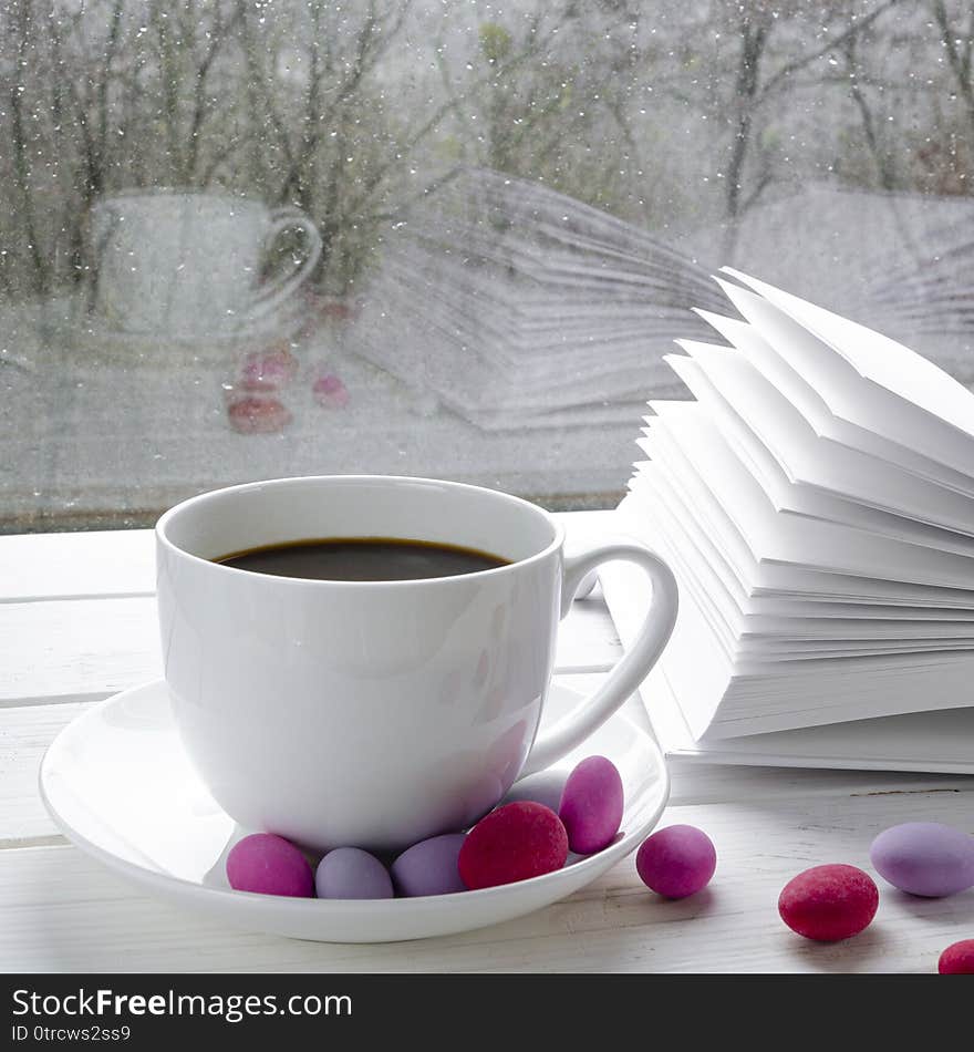 Cozy concept: a white cup of coffee stands on a white wooden table next to a white open book and colorful candy against the background of a window with raindrops and reflection on the glass, winter, snow, home, cold, warm, season, room, weather, house, tea, windowsill, calm, hot, weekend, comfort, gray, still, life, relax, rest, mood, ice, leisure, frame, blanket, copy, moody, drink, scarf, frost, retro, shabby, chic, view, indoors, interior, holiday, snowdrift, mug