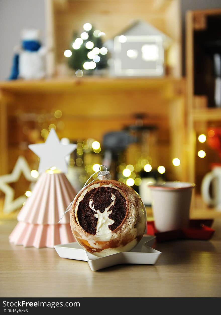 Christmas dessert. Tiramisu served in a glass bowl like a Christmas tree ball with deer image. Cup of coffee or tea. Ceramic Christmas tree with white star. Blurry lights and miniature house in the background. Christmas dessert. Tiramisu served in a glass bowl like a Christmas tree ball with deer image. Cup of coffee or tea. Ceramic Christmas tree with white star. Blurry lights and miniature house in the background