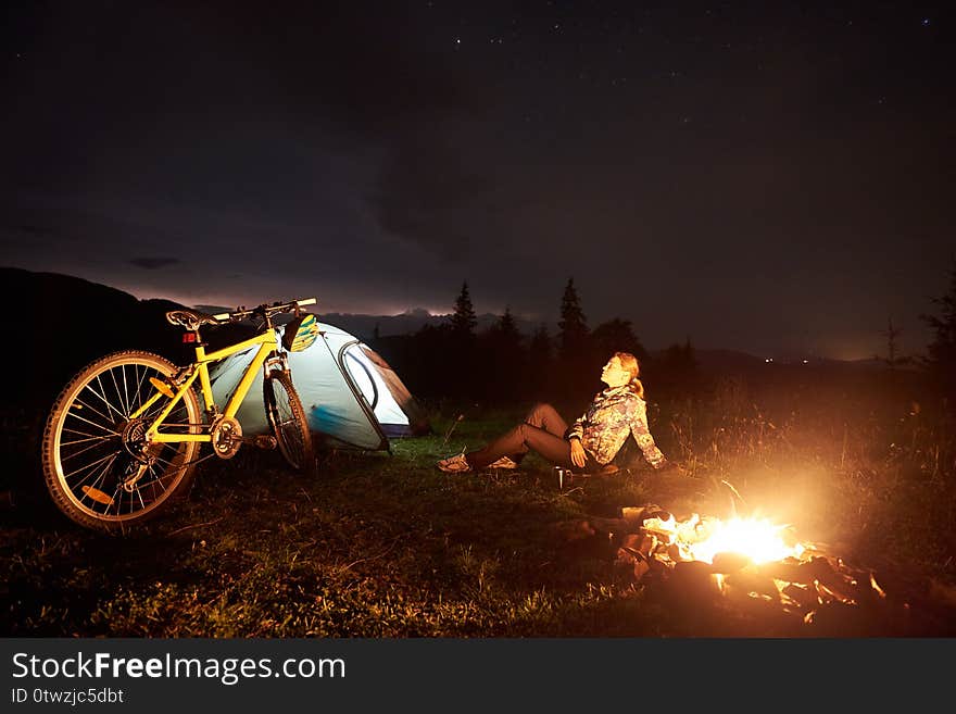 Woman resting at night camping near campfire, tourist tent, bicycle under evening sky full of stars